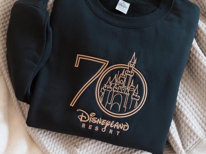 Puede incluir: Sudadera negra con dise&ntilde;o bordado en dorado. El dise&ntilde;o presenta el n&uacute;mero 70, un castillo dentro de un c&iacute;rculo y las palabras "Disneyland Resort". La sudadera est&aacute; doblada sobre una manta de color neutro.
