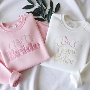 May include: Two crewneck sweatshirts, one pink and one white. The pink sweatshirt says "Bride" in embroidered letters, with a bow design above. The white sweatshirt says "Team Bride" in embroidered letters, with a bow design above.