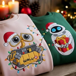 Puede incluir: Dos sudaderas con temática navideña. Una es de color rosa claro con un diseño de robot amarillo que lleva un gorro de Papá Noel y una guirnalda de luces. La otra es verde con un diseño de robot blanco que lleva un gorro de Papá Noel y un regalo.