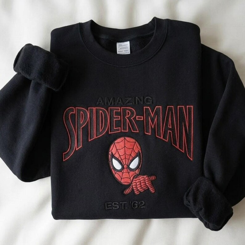 May include: A black long-sleeve sweatshirt featuring an embroidered red and black Spider-Man graphic. The text reads AMAZING SPIDER-MAN in red lettering above the character's head and EST. '62 in black thread below the character's hand.