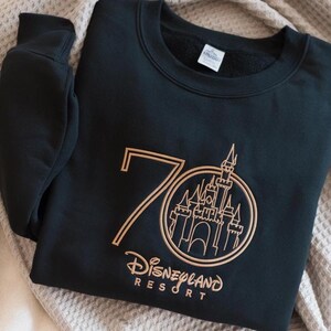 Puede incluir: Sudadera negra con un dise&ntilde;o bordado en dorado. El dise&ntilde;o incluye el n&uacute;mero 70, un castillo y las palabras "Disneyland Resort". La sudadera est&aacute; doblada sobre una superficie clara.