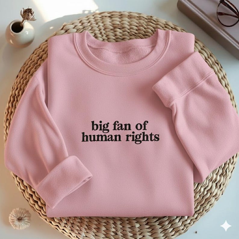 May include: A light pink sweatshirt with the text "big fan of human rights" in black. The sweatshirt is folded on a woven mat. The sleeves are cuffed. A small white vase with a dried flower and a pair of glasses are in the background.