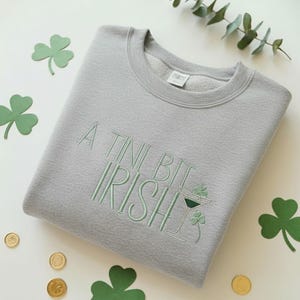 May include: A light gray sweatshirt with the embroidered phrase "A Tini Bit Irish" in light green. The design includes a martini glass with a shamrock and additional shamrocks. Gold coins and green shamrock cutouts are scattered around the sweatshirt.
