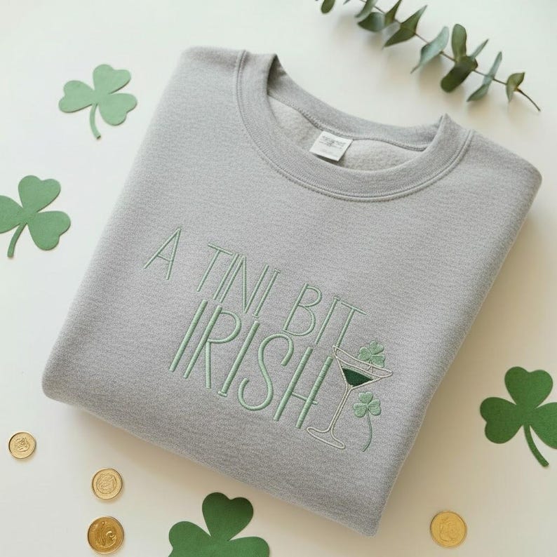 May include: A light gray sweatshirt with the embroidered phrase "A Tini Bit Irish" in light green. The design includes a martini glass with a shamrock and additional shamrocks. Gold coins and green shamrock cutouts are scattered around the sweatshirt.