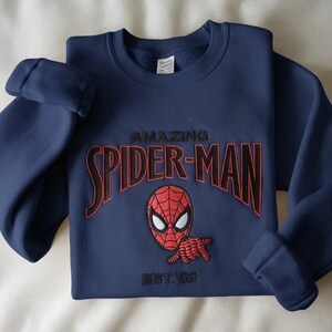 May include: A navy blue crewneck sweatshirt featuring an embroidered design of the Spider-Man mask and hand gesture. The text reads AMAZING SPIDER-MAN in black with red outlines, and EST. '62 is embroidered in black below the character graphic.