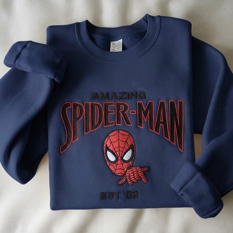 May include: A navy blue crewneck sweatshirt featuring an embroidered design of the Spider-Man mask and hand gesture. The text reads AMAZING SPIDER-MAN in black with red outlines, and EST. '62 is embroidered in black below the character graphic.