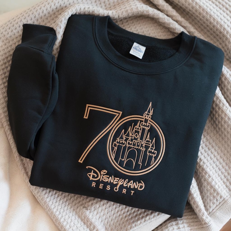 Puede incluir: Sudadera negra de cuello redondo con un dise&ntilde;o dorado del n&uacute;mero 70 y el castillo de Disneyland Resort. La sudadera est&aacute; doblada sobre una superficie texturizada de color neutro. El logotipo de Disneyland Resort est&aacute; debajo.