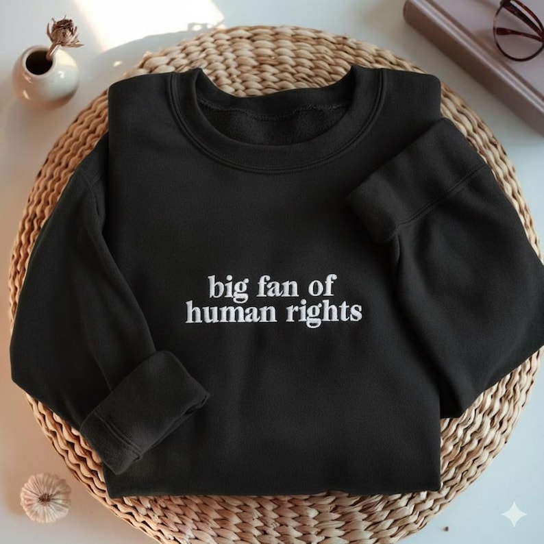 May include: Black sweatshirt with the embroidered text "big fan of human rights" in white. The sweatshirt is displayed on a woven, circular surface. The sleeves are cuffed, and the neckline is crew. A small vase with a flower is visible in the background.