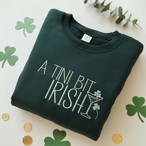 May include: Dark green sweatshirt with the embroidered phrase "A Tini Bit Irish" in light green. The design includes a martini glass with shamrocks. The sweatshirt is surrounded by shamrock cutouts and gold coins.