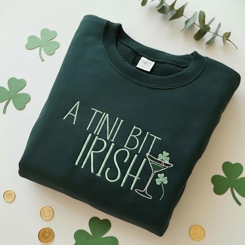 May include: Dark green sweatshirt with the embroidered phrase "A Tini Bit Irish" in light green. The design includes a martini glass with shamrocks. The sweatshirt is surrounded by shamrock cutouts and gold coins.