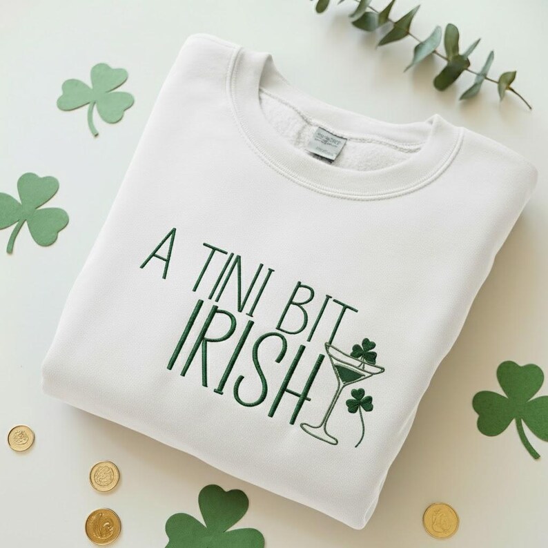 May include: White sweatshirt with the text "A Tini Bit Irish" in green, along with a martini glass and shamrock design. The sweatshirt is surrounded by shamrock cutouts and gold coins, perfect for St. Patrick's Day.
