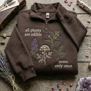 May include: A dark brown quarter-zip sweatshirt featuring embroidered botanical illustrations of various plants and flowers. The text "all plants are edible" is embroidered on the left, and "some only once" is embroidered on the lower right side.