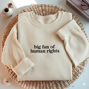May include: Cream-colored sweatshirt with the text "big fan of human rights" in black. The sweatshirt is folded on a woven, circular surface. A small vase with a dried flower and a book are in the background.