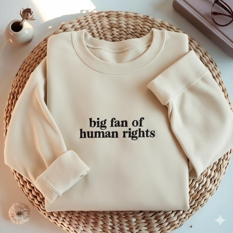 May include: Cream-colored sweatshirt with the text "big fan of human rights" in black. The sweatshirt is folded on a woven, circular surface. A small vase with a dried flower and a book are in the background.