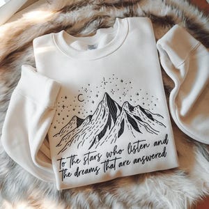May include: Cream-colored sweatshirt with a black mountain range design, stars, and a crescent moon. The text reads, "To the stars who listen and the dreams that are answered." The sweatshirt has long sleeves and a crew neck.
