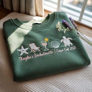 Puede incluir: Una sudadera verde oscuro con imágenes bordadas de una silla de playa, un cóctel, una concha, una estrella de mar y una tortuga marina. El texto "Kaylee's Bachelorette | Cape Cod 2025" está bordado debajo de las imágenes.