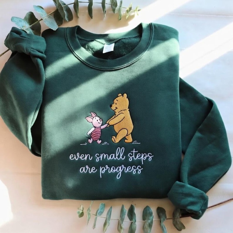 May include: A dark green sweatshirt featuring an embroidered design of Winnie the Pooh and Piglet holding hands. The text "even small steps are progress" is embroidered below the characters. The sweatshirt has long sleeves and a crew neck.