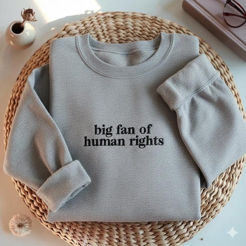 May include: A light gray sweatshirt with the text "big fan of human rights" in black. The sweatshirt is folded on a woven wicker mat. The sleeves are cuffed, and the neckline is crew-cut.