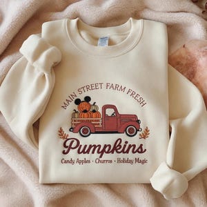 May include: A cream-colored crewneck sweatshirt featuring a graphic of a red pickup truck carrying pumpkins, one with mouse ears. Text reads: Main Street Farm Fresh Pumpkins, Candy Apples, Churros, Holiday Magic. The design is in brown and red tones.