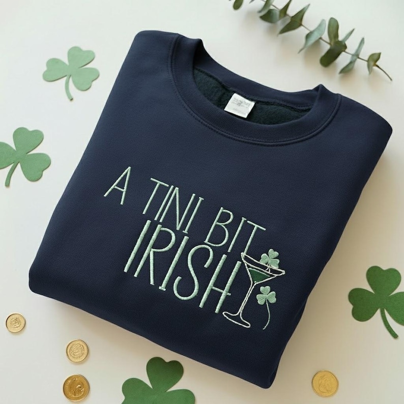 May include: Navy blue sweatshirt with the embroidered phrase "A Tini Bit Irish" in light green. The design includes a martini glass with shamrocks. The sweatshirt is surrounded by shamrock cutouts and gold coins.