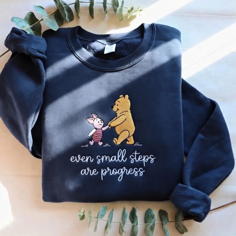 May include: Navy blue sweatshirt featuring an embroidered design of Winnie the Pooh and Piglet holding hands. The text "even small steps are progress" is written below the characters. The sweatshirt has long sleeves and a crew neck.