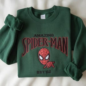 May include: A dark green crewneck sweatshirt featuring an embroidered graphic of the Spider-Man mask and hand gesture. The text reads AMAZING SPIDER-MAN in red and black lettering, with EST. '62 embroidered in black below the character graphic.
