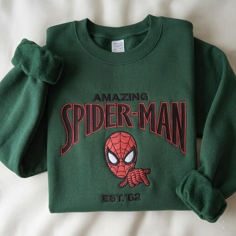 May include: A dark green crewneck sweatshirt featuring an embroidered graphic of the Spider-Man mask and hand gesture. The text reads AMAZING SPIDER-MAN in red and black lettering, with EST. '62 embroidered in black below the character graphic.