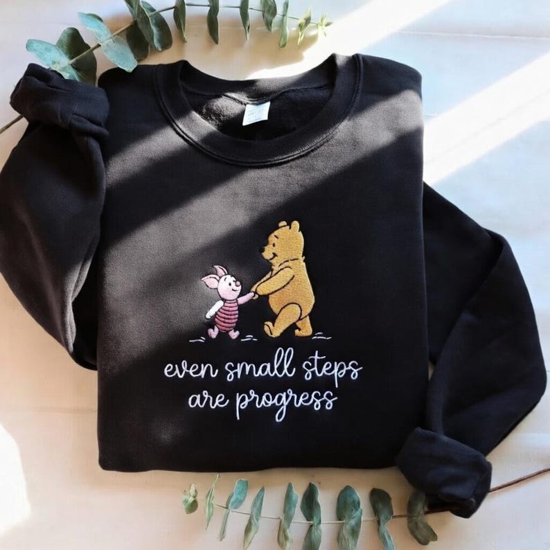 May include: Black sweatshirt featuring an embroidered design of Winnie the Pooh and Piglet holding hands. The text "even small steps are progress" is below the characters. The sweatshirt has long sleeves and a crew neck.