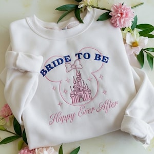 Puede incluir: Una sudadera blanca de cuello redondo con un dise&ntilde;o bordado de la silueta de un castillo rosa dentro del contorno de unas orejas de Mickey Mouse. El texto BRIDE TO BE est&aacute; bordado en azul arriba y Happy Ever After en cursiva rosa abajo.