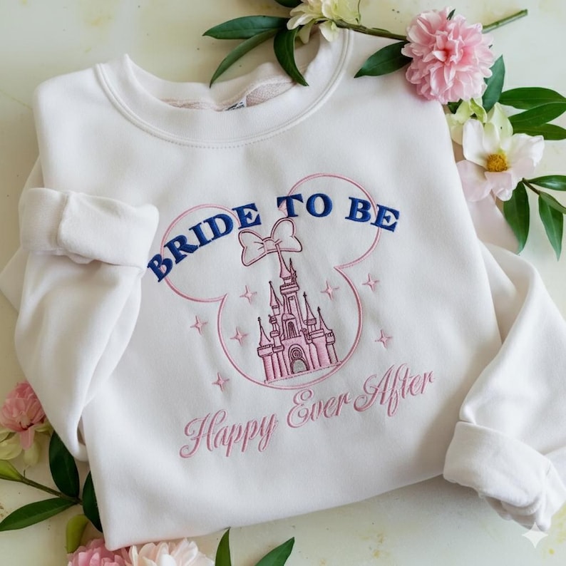 Puede incluir: Una sudadera blanca de cuello redondo con un dise&ntilde;o bordado de la silueta de un castillo rosa dentro del contorno de unas orejas de Mickey Mouse. El texto BRIDE TO BE est&aacute; bordado en azul arriba y Happy Ever After en cursiva rosa abajo.