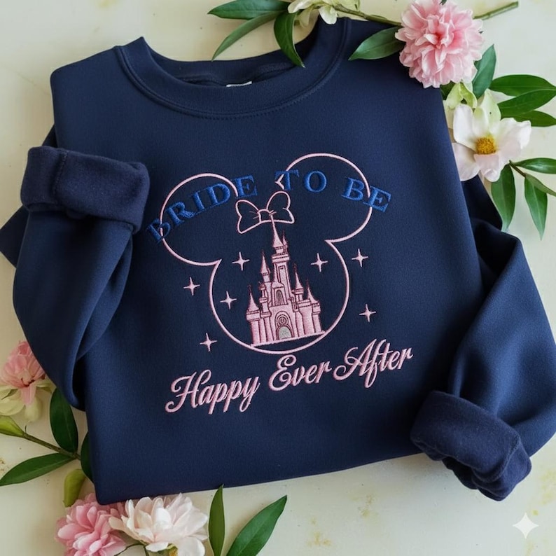 Puede incluir: Una sudadera azul marino de cuello redondo con un dise&ntilde;o bordado de la silueta de un castillo rosa dentro del contorno de unas orejas de Mickey Mouse. El texto BRIDE TO BE est&aacute; bordado en azul arriba y Happy Ever After en rosa abajo.