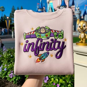 Puede incluir: Sudadera rosa claro con un diseño bordado. El diseño presenta las alas de Buzz Lightyear, la frase "to infinity" en escritura morada y un cohete. El diseño incluye estrellas amarillas. Cuello redondo.