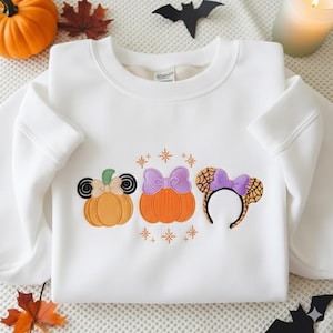 May include: A white crewneck sweatshirt featuring an embroidered design of two orange pumpkins and a pair of mouse ears with a spiderweb pattern. The pumpkins are adorned with purple and beige bows, surrounded by small embroidered starburst accents.