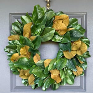 May include: A circular wreath composed of green and gold-colored leaves. The leaves are arranged in a dense, overlapping pattern, creating a full and textured appearance. The wreath is set against a gray door with a decorative frame and a gold-colored door knocker.