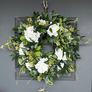 May include: A circular wreath featuring white hydrangea blooms, green leaves, and small green buds. The wreath is mounted on a gray door with a decorative gold-colored deer head hook.