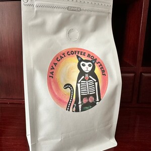 May include: White coffee bag with a zipper closure and a round vent. The bag features a colorful circular label with the text "JAVA CAT COFFEE ROASTERS" and an illustration of a cat skeleton.