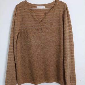 Puede incluir: Un suéter marrón de manga larga con cuello henley y tres botones. El suéter tiene un aspecto texturizado y está colgado de una percha blanca sobre un fondo blanco. Las mangas tienen un patrón de rayas.