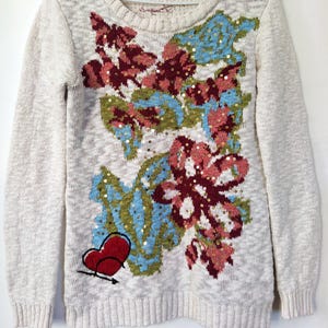 May include: A cream-colored sweater with a floral design featuring sequins and a red heart. The sweater has long sleeves and a crew neck.