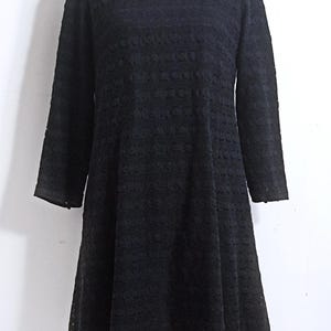 May include: A black, long-sleeved dress with a round neckline. The dress features a textured, openwork design throughout the fabric. The hemline is slightly flared, and the sleeves extend to the wrists. The dress is displayed on a white mannequin.