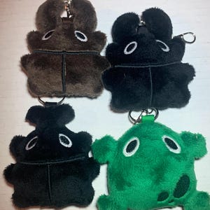May include: Four plush keychains in the shape of cartoon characters. The keychains are brown, black, black, and green. The characters have large eyes and a simple design.