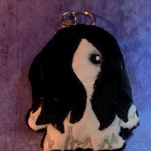 May include: A plush keychain in the shape of a black and white dog. The dog has floppy ears and a small, round eye. The keychain ring is silver. The dog's fur is black and white, with a soft, plush texture.