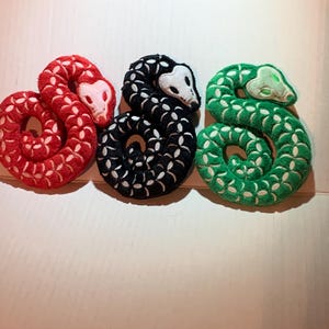 May include: Three coiled snake-shaped plush toys in red, black, and green. Each snake has a white face and a pattern of white floral shapes. The red snake has a white face, the black snake has a white face, and the green snake has a light green face.