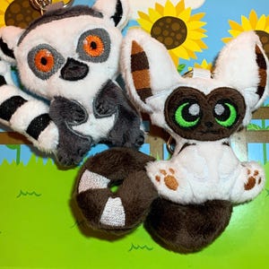 May include: Two plush animal keychains. One is a lemur with a black and white striped tail, gray arms, and orange eyes. The other is a white and brown creature with green eyes and a brown tail. The background features sunflowers.