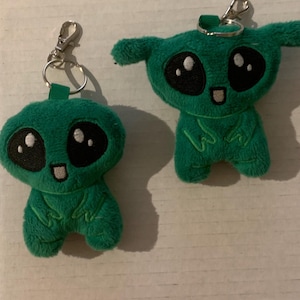May include: Two green plush alien keychain toys with big eyes and a smiling mouth. The larger alien has a key ring attached to its head.