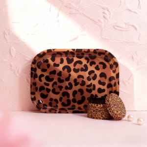 May include: A rectangular serving tray and a small round container with a lid, both featuring a brown and black leopard print pattern. The tray has rounded corners. The items are on a pink surface.