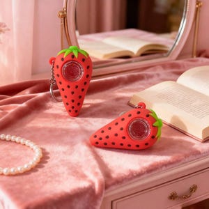 May include: Two red strawberry-shaped keychains with black seeds and green tops are displayed on a pink surface. The keychains have a clear center. One keychain is upright, and the other is lying on its side.