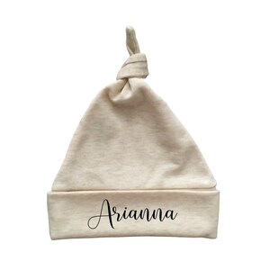 May include: A beige baby knot hat with the name "Arianna" in black script across the folded brim. The hat is made of soft fabric and has a knotted top.