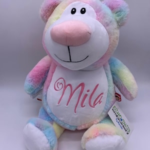 May include: A plush rainbow-colored bear with the name "Mila" embroidered in pink on its chest. The bear has a white face, pink nose, and black eyes. A tag from "Echo House Embroidery" is attached.