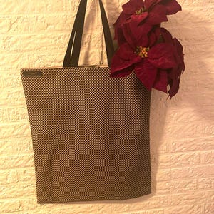 Op de afbeelding: Een bruin-zwarte geruite draagtas met zwarte handvatten hangt tegen een witte bakstenen muur. De tas bevat dieprode poinsettia-bloemen. Een stijlvol en functioneel accessoire.