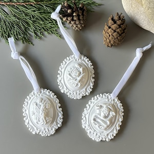 May include: Three white oval ornaments featuring intricate relief designs of a figure, a flower, and a skull with a snake. Each piece is framed by a decorative border and includes a white ribbon loop for hanging, providing a textured appearance.